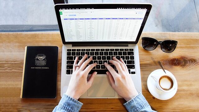 ノートパソコンに向かって作業をする手元、脇にノートとコーヒー