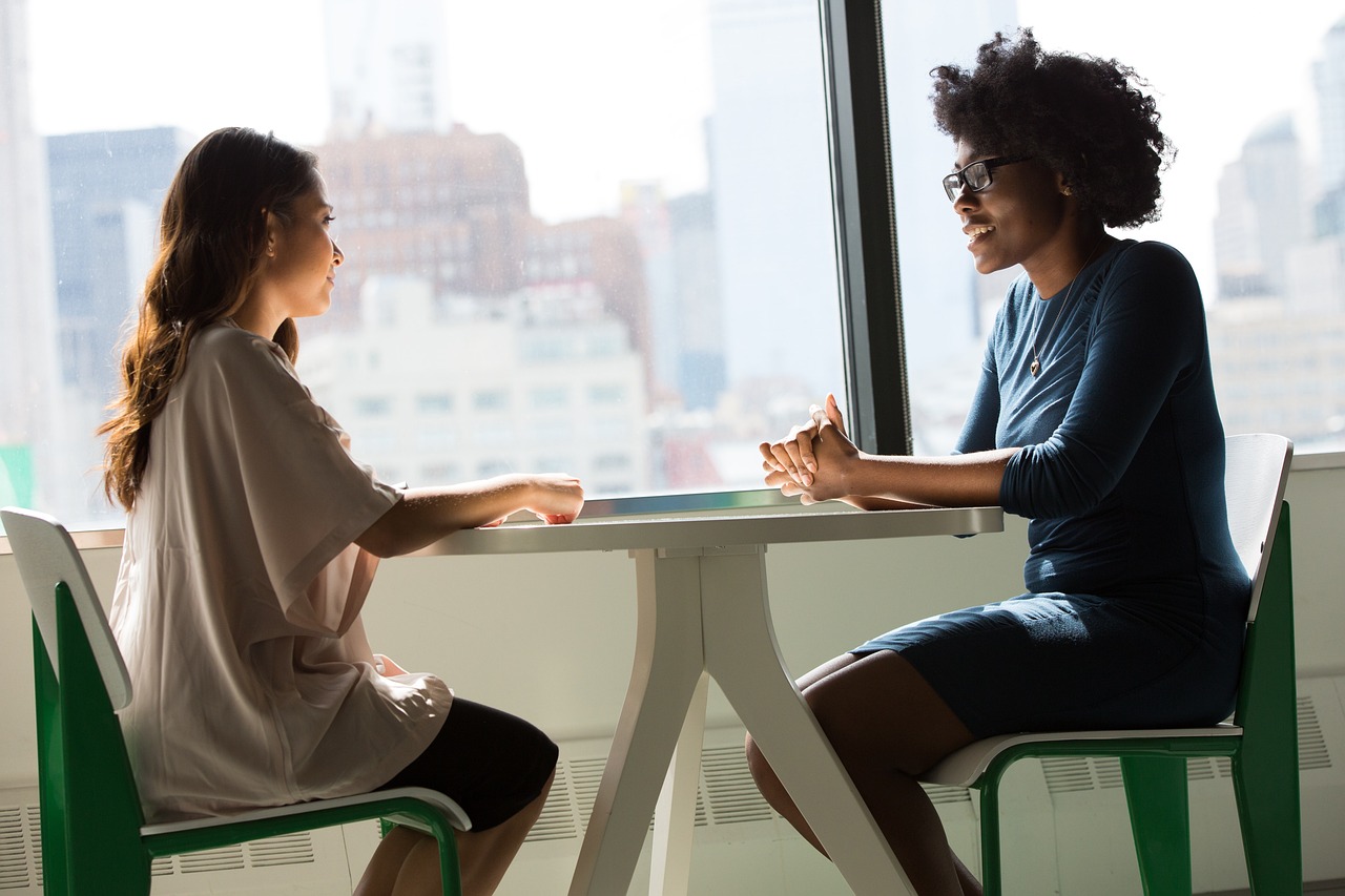 オフィスの窓際のテーブルで向かい合い、笑顔で会話している二人の女性