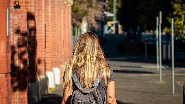 リュックを背負い、日差しの差し込む道を歩く女性の後ろ姿