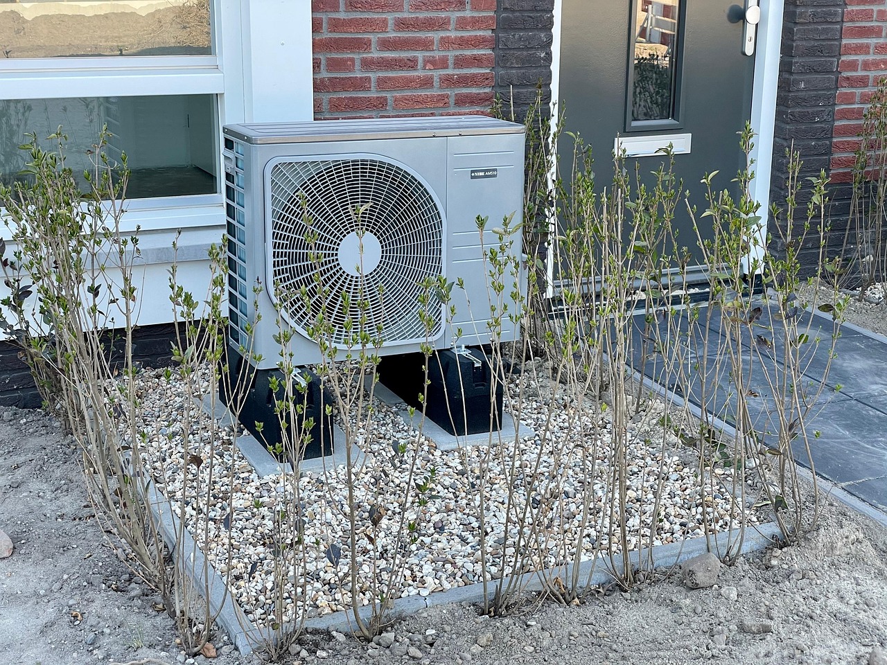 家の前の砂利の上に置かれ、まわりに植え込みがある室外機