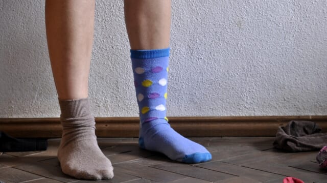 壁の前に立つ人の脚。左右で違う柄の靴下をはき、床にも靴下が散らばっている