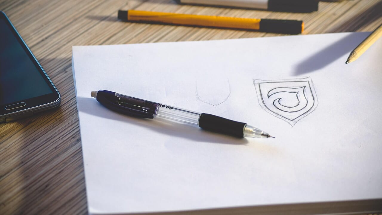 スケッチブックに鉛筆で描かれた盾形のロゴの下書きとシャープペンシル
