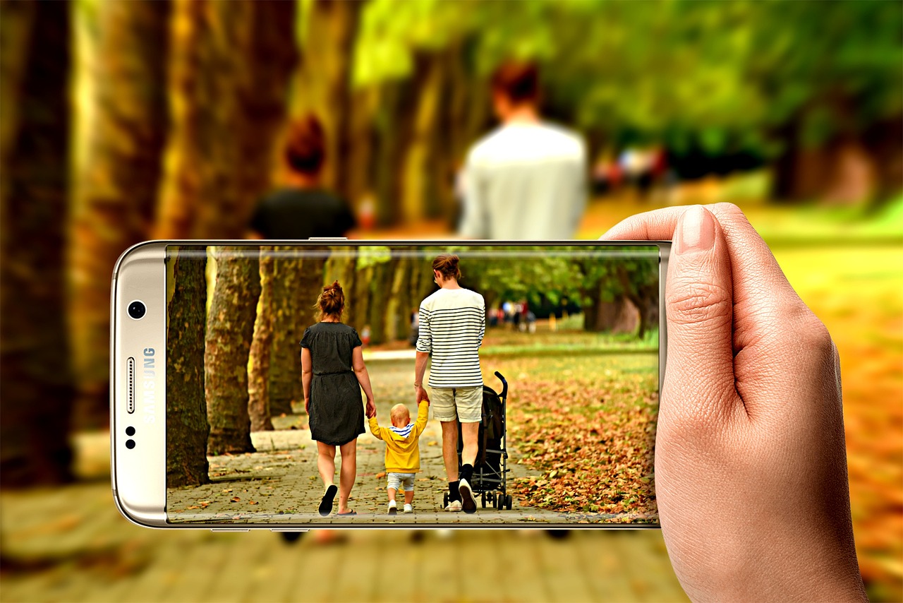 公園の並木道を歩く家族の後ろ姿を、手に持ったスマートフォンのカメラで写している様子