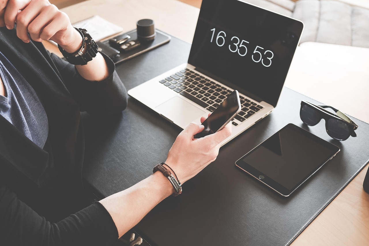 ノートパソコンとタブレットを並べた机で、スマートフォンを操作する人