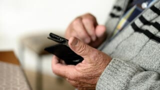 灰色のカーディガンを着た年配の人が、黒いスマートフォンを手に持っている手元