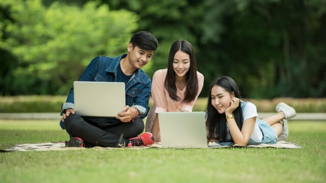 芝生の上でノートパソコンを見ながら笑う3人の学生