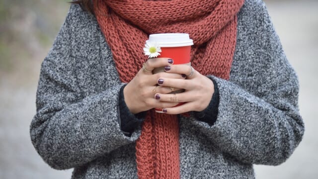 コートとマフラー姿で、両手で温かい飲み物のカップを持つ人