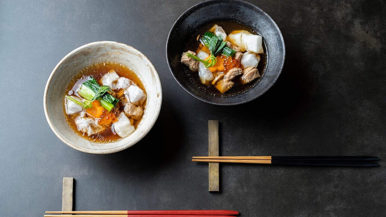 黒い器と白い器に入った2杯の雑煮。餅と青菜、鶏肉が入り、赤い箸と黒い箸が添えられている