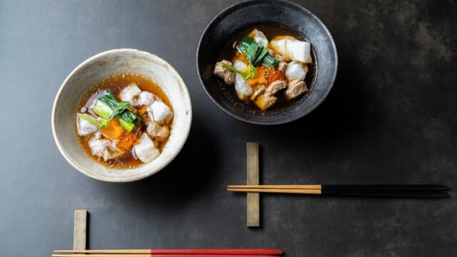 黒い器と白い器に入った2杯の雑煮。餅と青菜、鶏肉が入り、赤い箸と黒い箸が添えられている