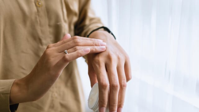 白いクリームの容器を持ち、手の甲に保湿クリームを塗っている手元