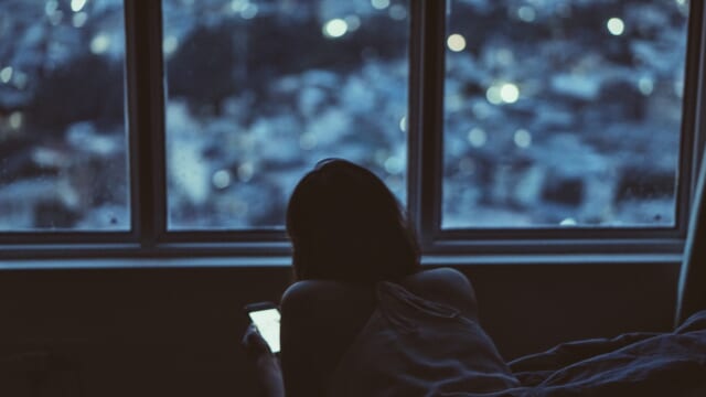 夜の暗い部屋で、窓辺のベッドに寝そべってスマホを見る女性の後ろ姿