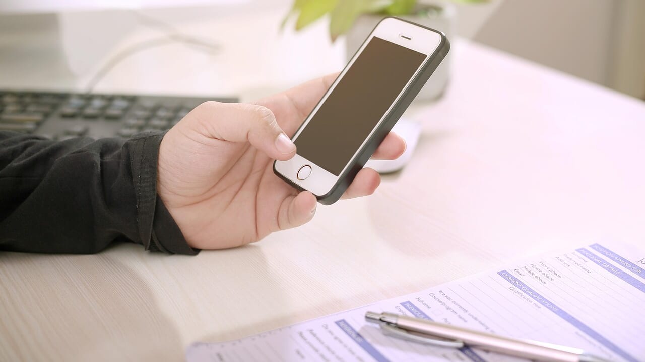 白い机の上で、書類とペンのそばにiPhoneを持っている手元