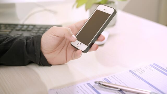 白い机の上で、書類とペンのそばにiPhoneを持っている手元