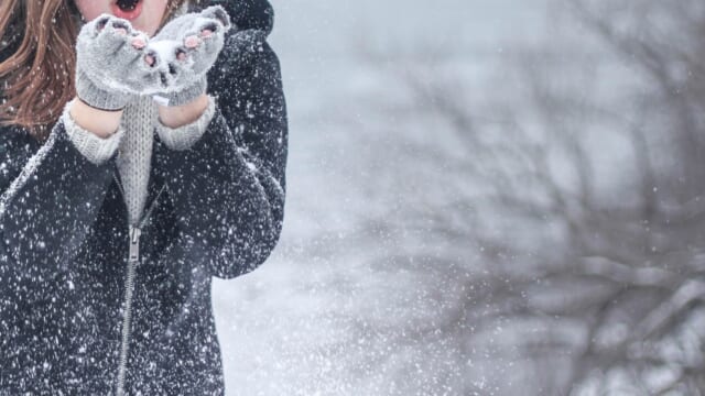 雪が舞う冬の屋外で、コートと手袋を身につけて手に息を吹きかける女性